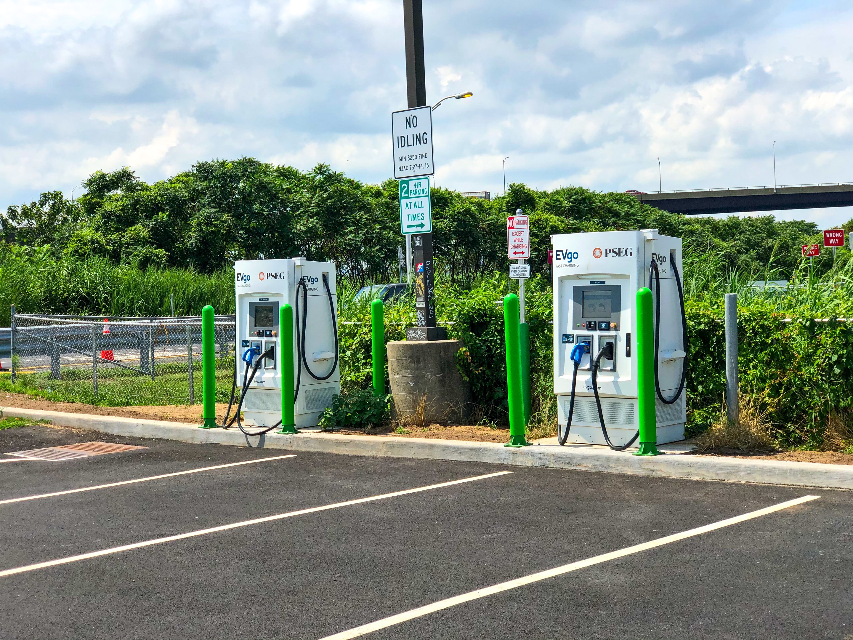 Elektrofahrzeug-Ladestation auf einem Parkplatz mit umgeben-der Infrastruktur, Grünflächen und einer Brücke im Hintergrund.
