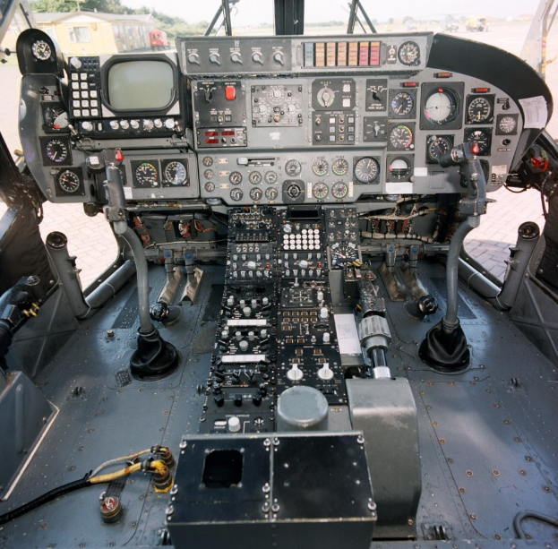 Ein Kampfflugzeugcockpit mit verschiedenen Instrumenten und Messgeräten, mit Bäumen, Gebäuden und einem klaren blauen Himmel im Hintergrund.