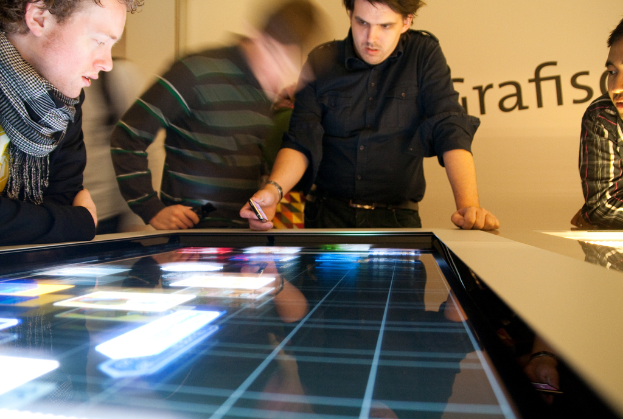 Spieler Spielen Fußball Auf Einem Feld Mit Einem Tor, Während Zuschauer Dahinter Stehen, Mit Bäumen Und Häusern Im Hintergrund.
