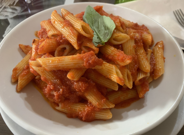 Ein weißer Teller mit Penne-Nudeln in Tomatensoße, garniert mit frischen Basilikumblättern, mit einer Gabel und einem Messer daneben.
