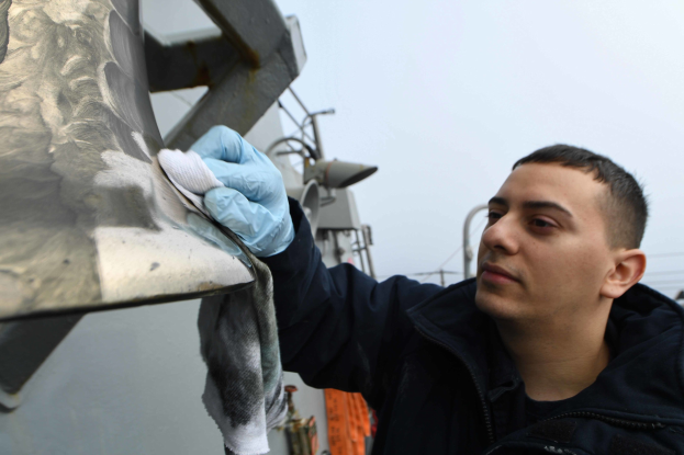 Ein Mann in einer schwarzen Jacke und blauen Handschuhen reinigt ein Metallobjekt mit einem Tuch im Freien, mit Strommasten, Drähten und einem klaren blauen Himmel im Hintergrund.