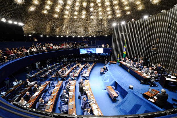 Eine Konferenzbühne mit sitzenden Personen, einem Tisch mit Papieren und Gegenständen, Flaggen und einer Skulptur im Hintergrund, mit Sitzplätzen an Tischen und hinter einer Absperrung, beleuchtet von Deckenlampen.