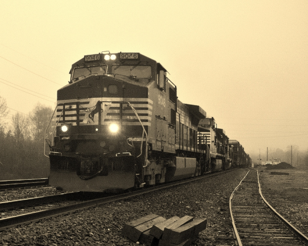 Ein Zug auf Eisenbahnschienen mit Autos, Strommasten, Bäumen und Himmel im Hintergrund.