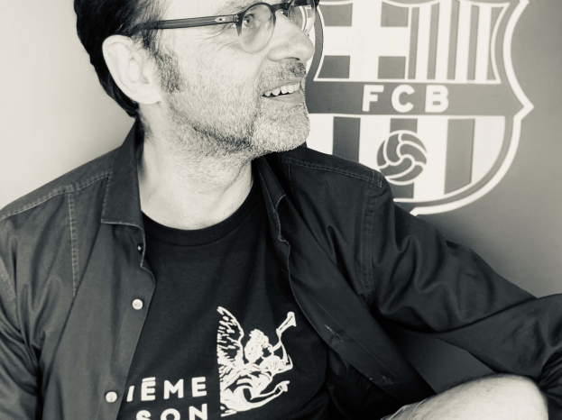 Schwarzes und weißes Foto eines grinsenden Mannes in einer Brille und einem schwarzen Hemd, der vor dem FC Barcelona-Logo sitzt.