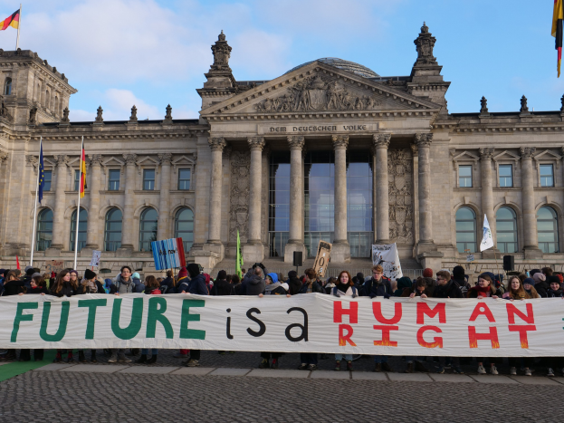 Eine Gruppe von Menschen mit einem Transparent mit der Aufschrift 'Zukunft ist ein Menschenrecht' vor dem Reichstaggebäude in Berlin, Deutschland, mit sichtbaren Fenstern, Säulen und Bögen, umgeben von Fahnenmasten, unter einem bewölkten Himmel.