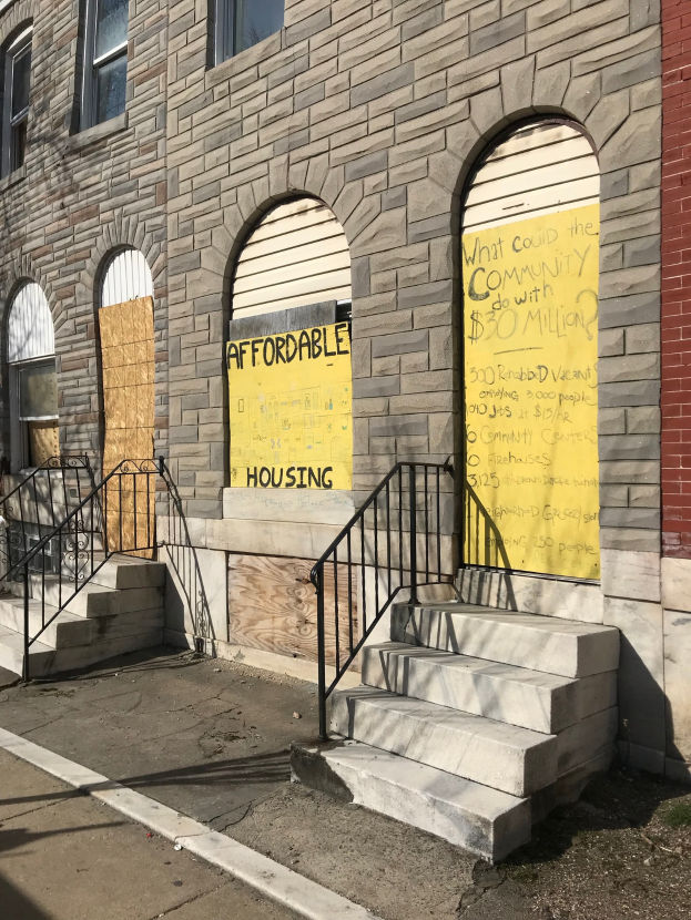 Ein zweistöckiges Gebäude mit vernagelten Fenstern und einem Schild mit der Aufschrift "Bezahlbarer Wohnraum", das über eine Treppe mit Geländern erreichbar ist und neben einer Straße steht.