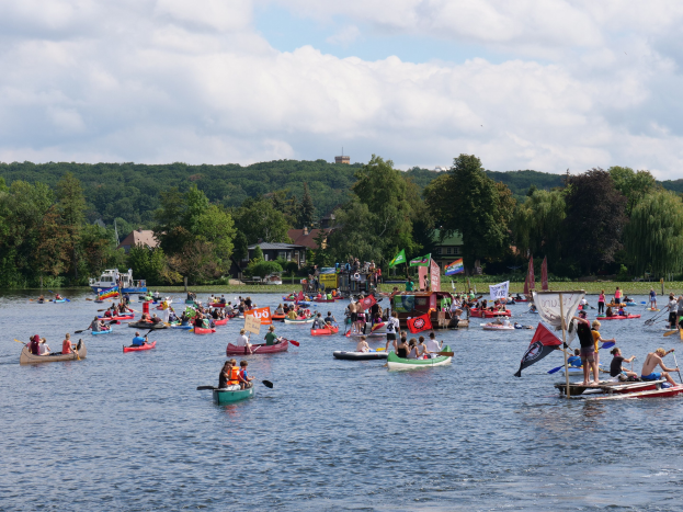 Eine große Gruppe von Menschen in Kanus auf einem Gewässer während eines Kanurennens, einige halten Paddel und Fahnen, vor dem Hintergrund von Bäumen, Gebäuden und einem bewölkten Himmel.