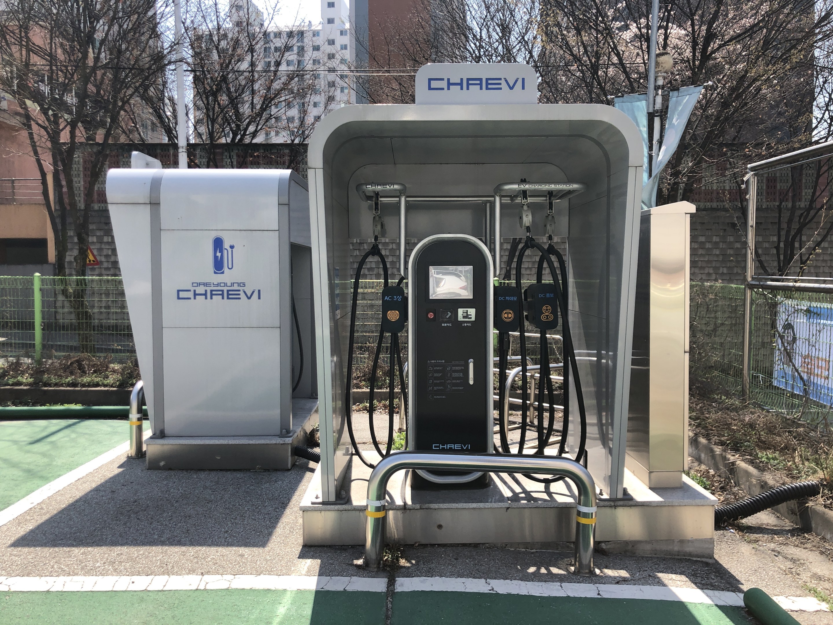 Elektroauto-Ladestation in einem Park, umgeben von einem Metallzaun, mit einer Fahne, Masten, Bäumen, Pflanzen, Gras, Gebäuden und einem bewölktem Himmel.