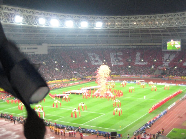 Ein Stadion mit Flutlicht, Eisengittern, Zuschauern, stehenden Personen, Werbetafeln, Anzeigetafeln, Überwachungskamera und verschiedenen Dekorationen einschließlich Flaggen.