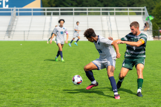 Eine Gruppe von Männern, die auf einem Rasenfeld umgeben von Bäumen Fußball spielen, mit Treppen, Geländern und einer Tafel mit Text im Hintergrund.