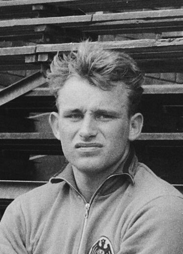 Schwarzes und weißes Foto eines Mannes in einer Jacke, der vor gestapelten Holzlatten steht, der angeblich ein legendärer deutscher Fußballspieler ist.