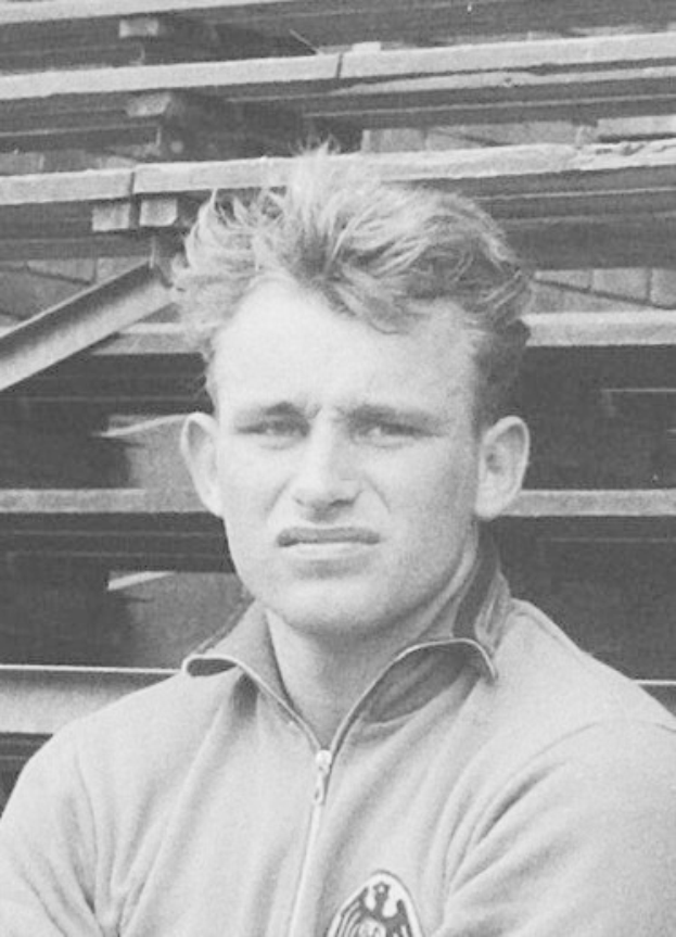 Schwarzes und weißes Foto eines Mannes in einer Jacke, der vor gestapelten Holzlatten steht, der angeblich ein legendärer deutscher Fußballspieler ist.