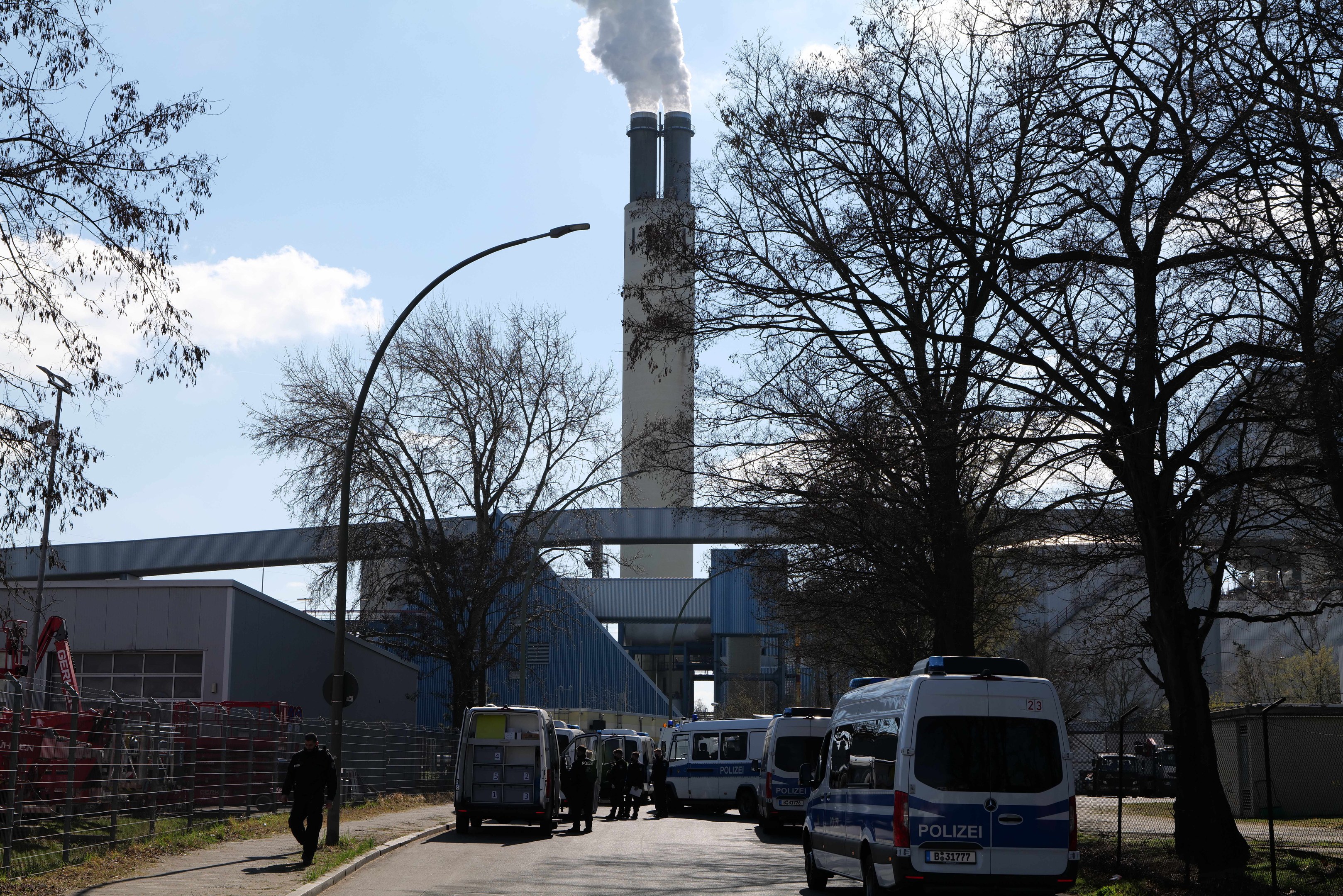 Gruppe von Polizeiwagen vor einem Fabrikgebäude mit Rauch aus dem Schornstein geparkt, mit Menschen auf der Straße und Gebäuden, Bäumen und Laternen im Hintergrund unter einem bewölkten Himmel.