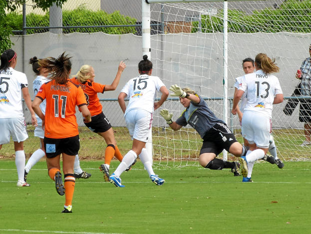 Eine Gruppe von Frauen spielt ein Spiel auf dem Boden mit einem Maschendrahtzaun und Bäumen im Hintergrund.