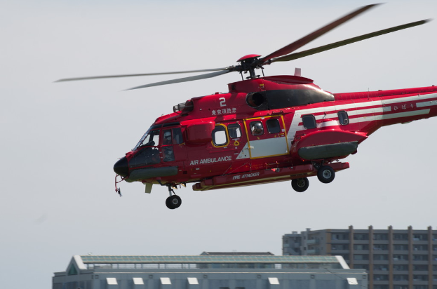 Ein roter Hubschrauber fliegt links mit Gebäuden unten und einem Himmel im Hintergrund.