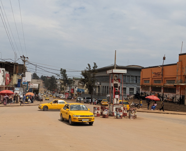 Ein gelbes Taxi fährt auf einer belebten Stadtstraße mit Gebäuden an der Seite, während Fußgänger mit Schirmen auf dem Gehweg unter bewölktem Himmel gehen, mit Strommasten und Kabeln im Hintergrund.