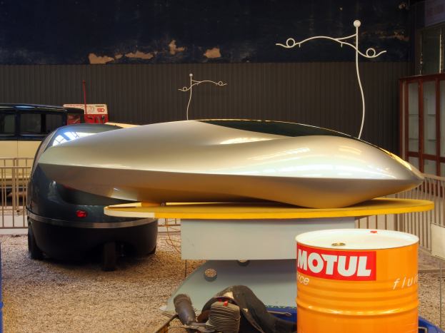 Kleines Hydrofoil-Boot auf einem Tisch neben einem Auto, mit zwei Trommeln auf dem Boden, einem Metallzaun und einer Glastür mit Text auf einer Tafel im Hintergrund.