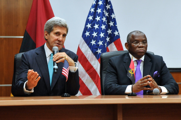 Zwei Männer in Blazern, Krawatten und Brillen sitzen an einem Tisch mit Mikrofonen, mit Flaggen dahinter, was auf eine formelle Diskussion hindeutet.