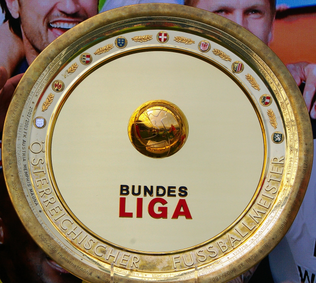 Ein Mann hält ein goldenes Schild mit 'Bundesliga' darauf, im Hintergrund eine Fahne mit drei Personen.