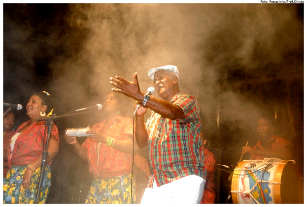 Eine Gruppe von Musikern, die auf der Bühne stehen, mit einem Mann, der in der Mitte in ein Mikrofon singt und einem anderen Mann, der rechts ein Schlagzeug spielt.