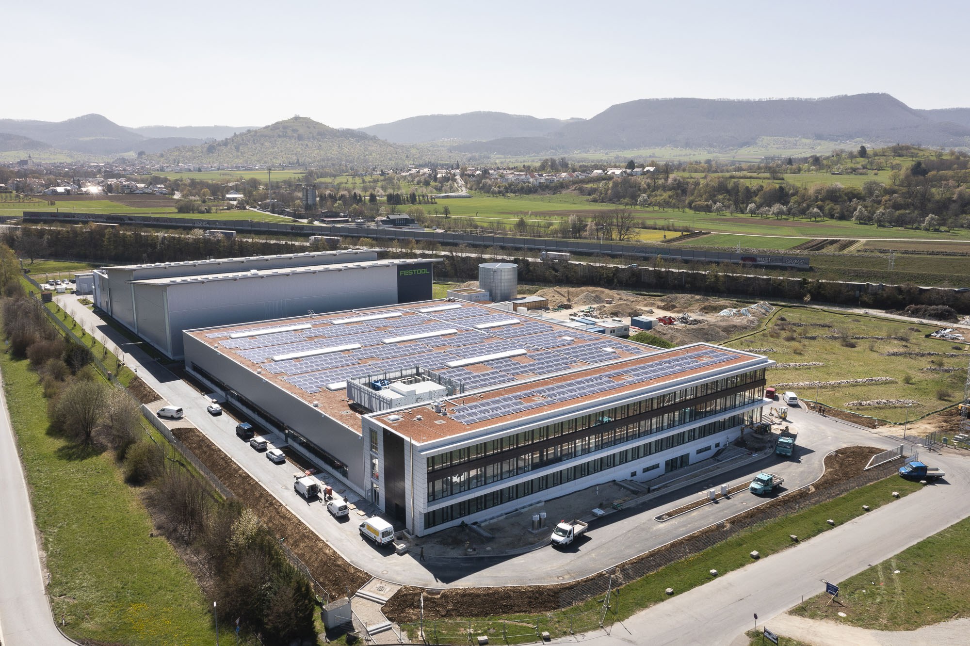 Luftaufnahme eines großen Industriegebäudes mit Solarmodulen auf dem Dach, umgeben von Fahrzeugen, Bäumen, Gras und Strommasten, mit Hügeln und einem klaren blauen Himmel im Hintergrund.
