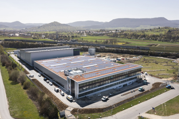 Luftaufnahme eines großen Industriegebäudes mit Solarmodulen auf dem Dach, umgeben von Fahrzeugen, Bäumen, Gras und Strommasten, mit Hügeln und einem klaren blauen Himmel im Hintergrund.