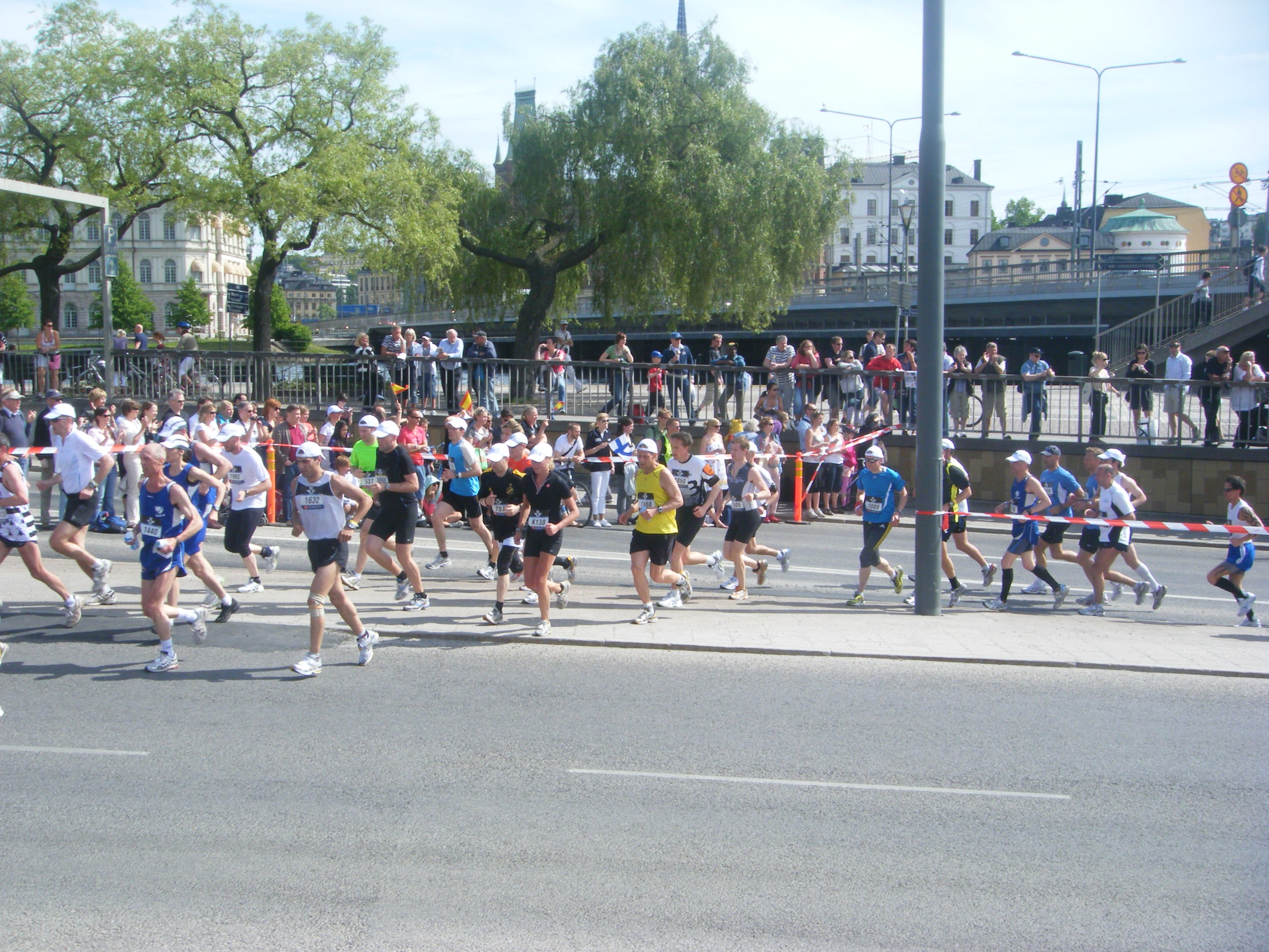 Gruppe von Menschen, die bei einem Marathon auf einer Straße mit Absperrungen, Zuschauern und einer Ziellinienflagge laufen.