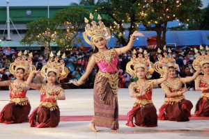Eine Gruppe von Frauen in traditioneller Thai-Tracht tanzt vor einem sitzenden und stehenden Publikum im Freien, mit Bäumen, Lichtern, Pfählen, einem Gebäude und einem klaren blauen Himmel im Hintergrund.