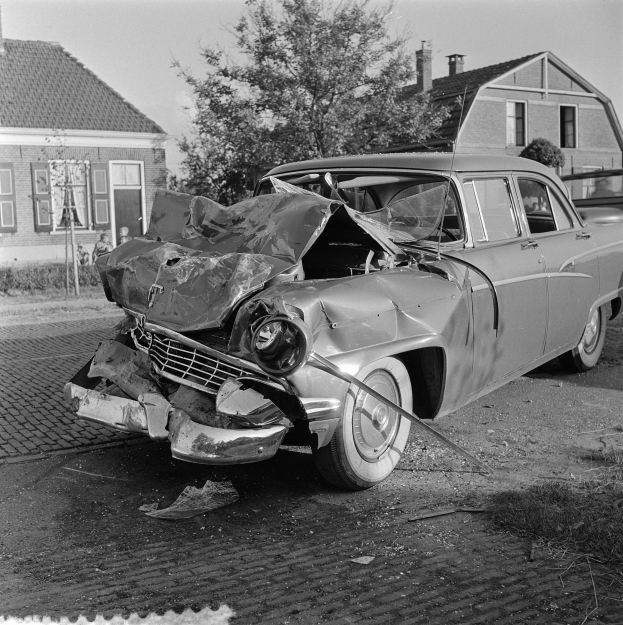 Ein Schwarz-Weiß-Foto eines schwer beschädigten Autos am Straßenrand mit eingedellter Motorhaube und zersplitterten Fenstern vor einer Kulisse aus Gebäuden, Bäumen und einem klaren Himmel.