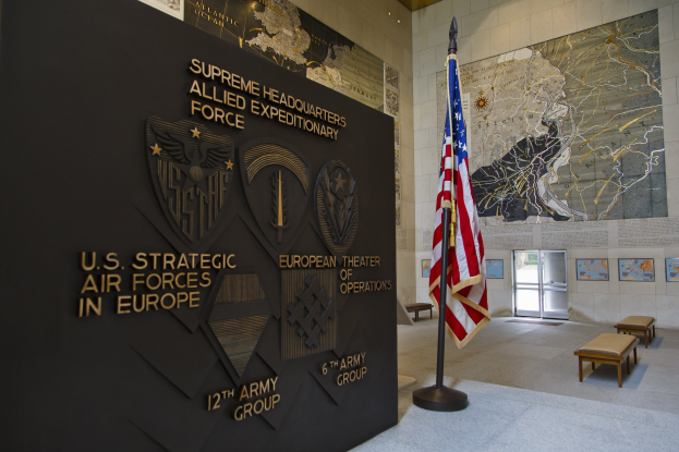 Eingang zum US-Strategischen Luftstreitkräfte-Hauptquartier in Europa mit einer amerikanischen Flagge, einer Wand mit Text und Logos, Bänken, gerahmten Fotos, einer Texttafel, beleuchteter Decke und einer Tür im Hintergrund.