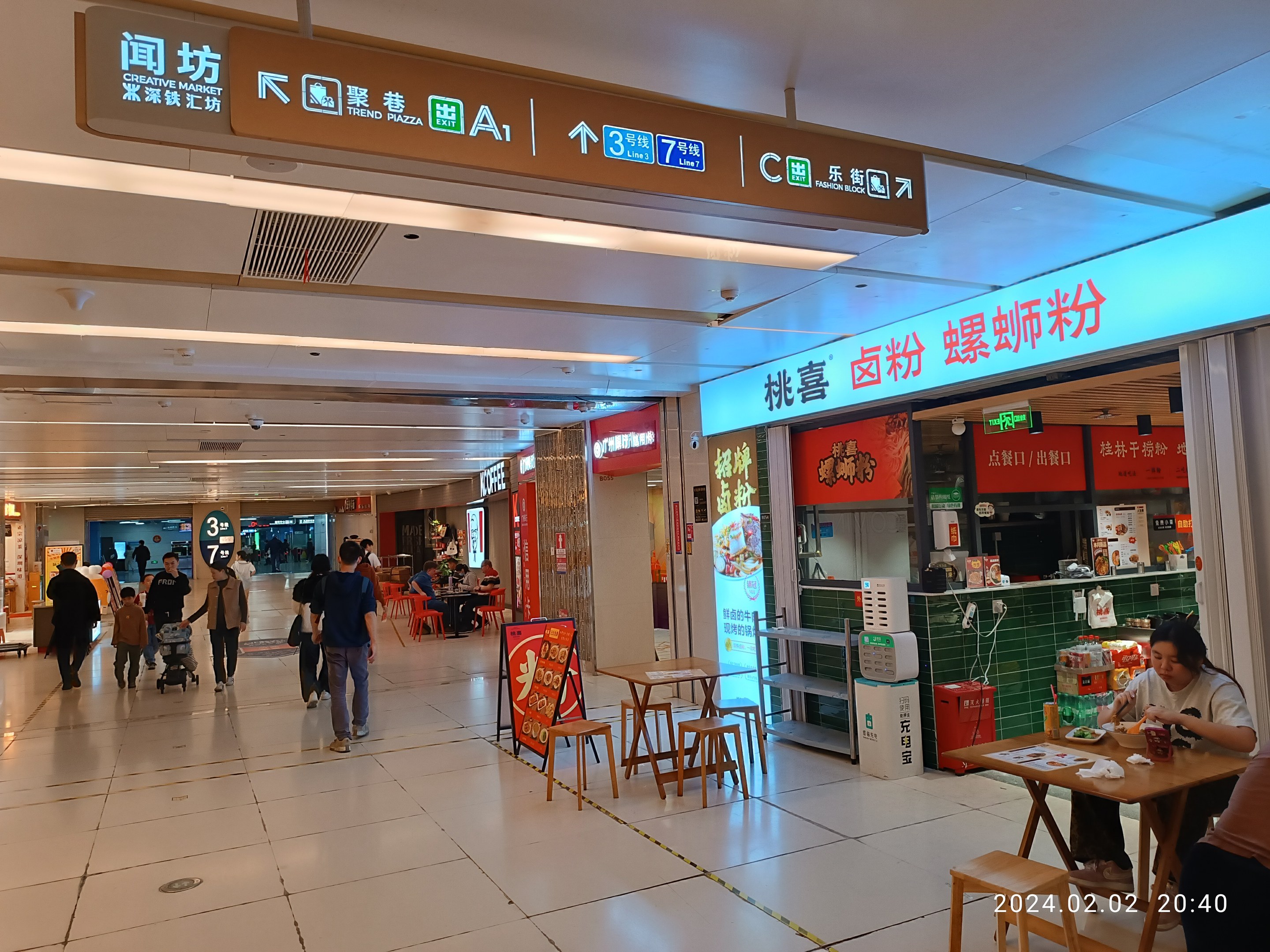 Menschen gehen durch ein Einkaufszentrum mit Tischen, Stühlen und einem Markt, der Lebensmittel anbietet, während Schilder mit Text an den Wänden angebracht sind.