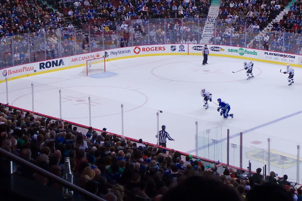 Vier Männer spielen Hockey auf einem Eisstadion, zwei stehen in der Nähe des Tors und des Zauns, Zuschauer sitzen auf Stühlen im Vorder- und Hintergrund, sowie eine Treppe im Hintergrund zu sehen.