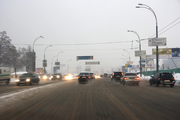 Eine städtische Straße voller Verkehr an einem verschneiten Tag, mit Fahrzeugen auf der Straße, schneebedeckter Erde, Laternen, Texttafeln, Bäumen, Gebäuden und einem Himmel im Hintergrund.