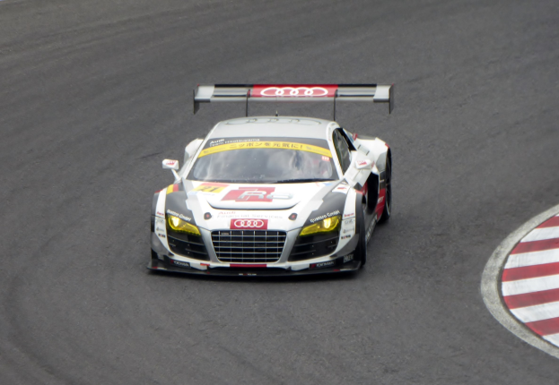 Ein leuchtend roter und weißer Audi R8 LMS GT3 Rennauto mit dem Audi-Logo auf der Seite, gefahren von einer Person in einem schwarzen Rennanzug und Helm, der eine Rennstrecke umfährt.