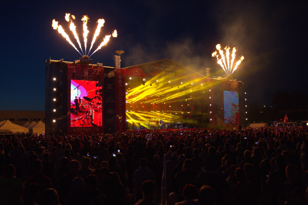 Eine große Menschenmenge auf einem Musikfestival mit einer Bühne, auf der Künstler unter hellen Scheinwerfern spielen, umgeben von Bannern, Zelten links und Feuerwerk am dunklen Himmel.