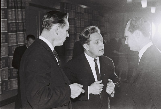 Drei Männer in Anzügen unterhalten sich in einem schwach beleuchteten Raum mit Bücherregalen im Hintergrund, aufgenommen in Schwarz-Weiß.