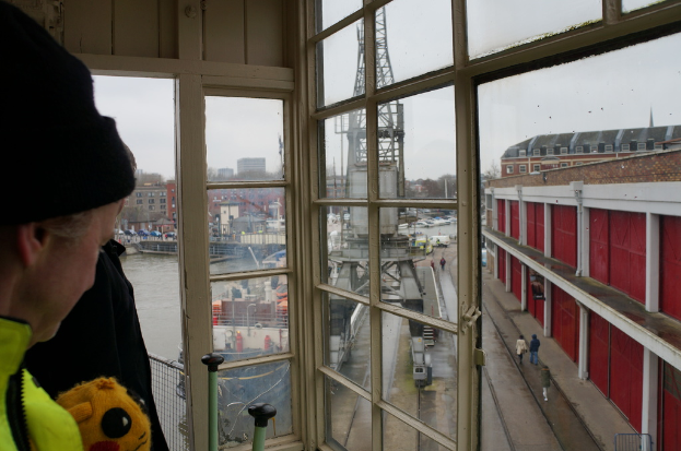 Eine Person bedient einen Turmdrehkran mit Gebäuden und Wasser im Vordergrund.