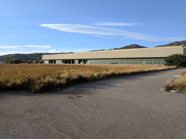 Leere Straße, die zu einem großen Gebäude in einem Feld führt, umgeben von Pflanzen und Bäumen, mit Hügeln und einem bewölkten Himmel im Hintergrund; das Gebäude ist die San Francisco International Airport-Zentrale.