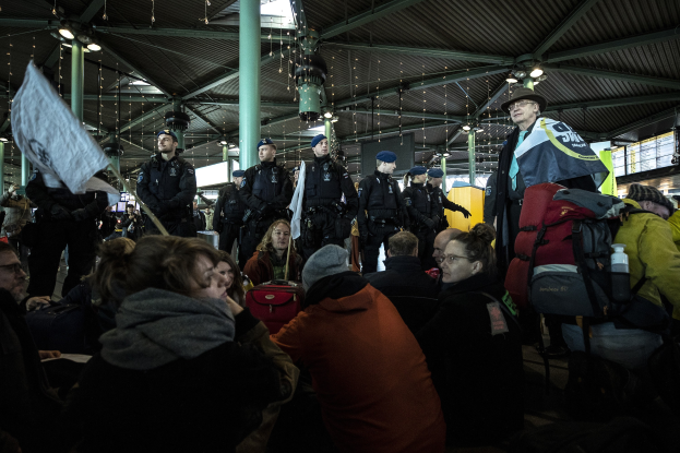 Eine Gruppe von Menschen steht vor einer Menge in einem Bahnhof, einige halten Schilder und Banner, andere tragen Mützen und tragen Taschen, sowie Säulen und Deckenleuchten im Hintergrund.