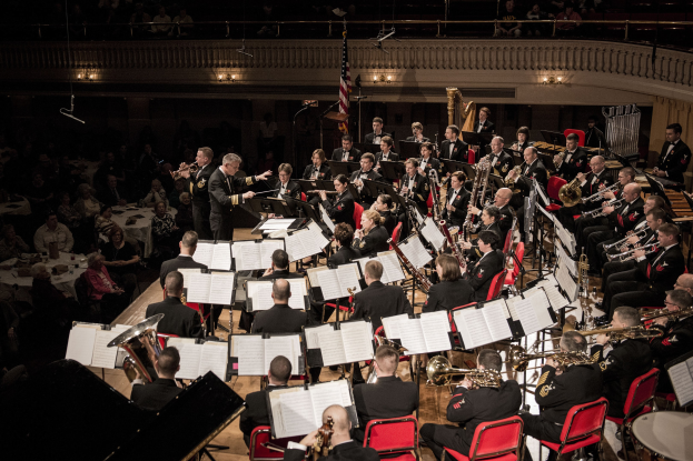 Eine Militärmusik in Uniform spielt in einem großen Raum mit sitzenden und stehenden Musikern, Notenpulten und einer Flagge an der Rückwand.