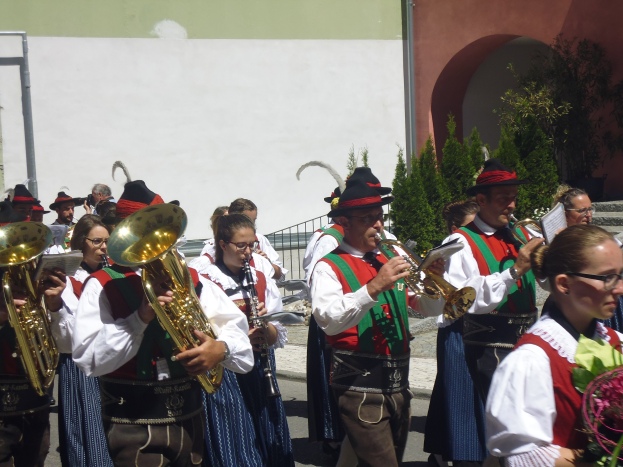 Eine Gruppe von Menschen in traditioneller bayerischer Tracht spielt Posaunen vor einem Gebäude, umgeben von Pflanzen, Geländern und einer Wand, wobei einige eine Brille tragen und musikalische Instrumente oder Blumen halten.