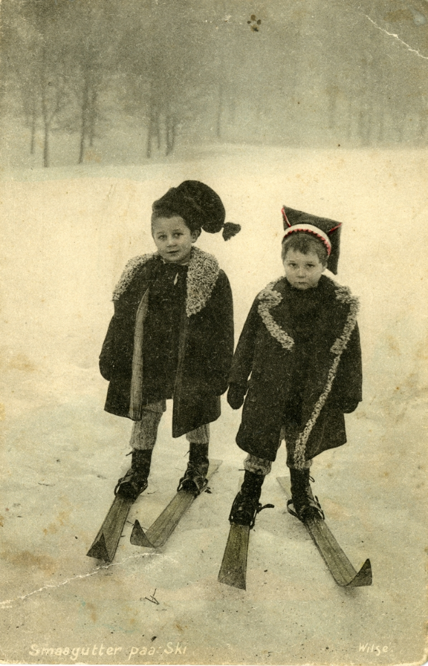 Zwei Kinder auf Skiern im Schnee, umgeben von Bäumen im Hintergrund, stehen auf schneebedeckter Erde.