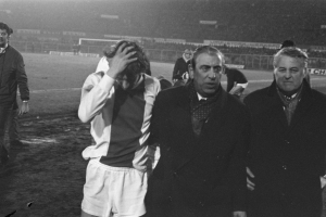 Schwarzes und weißes Foto einer Gruppe von Männern auf einem Fußballfeld, die nach einem Spiel enttäuscht aussehen, mit einem Stadion im Hintergrund.