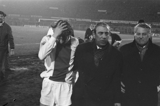 Schwarzes und weißes Foto einer Gruppe von Männern auf einem Fußballfeld, die nach einem Spiel enttäuscht aussehen, mit einem Stadion im Hintergrund.