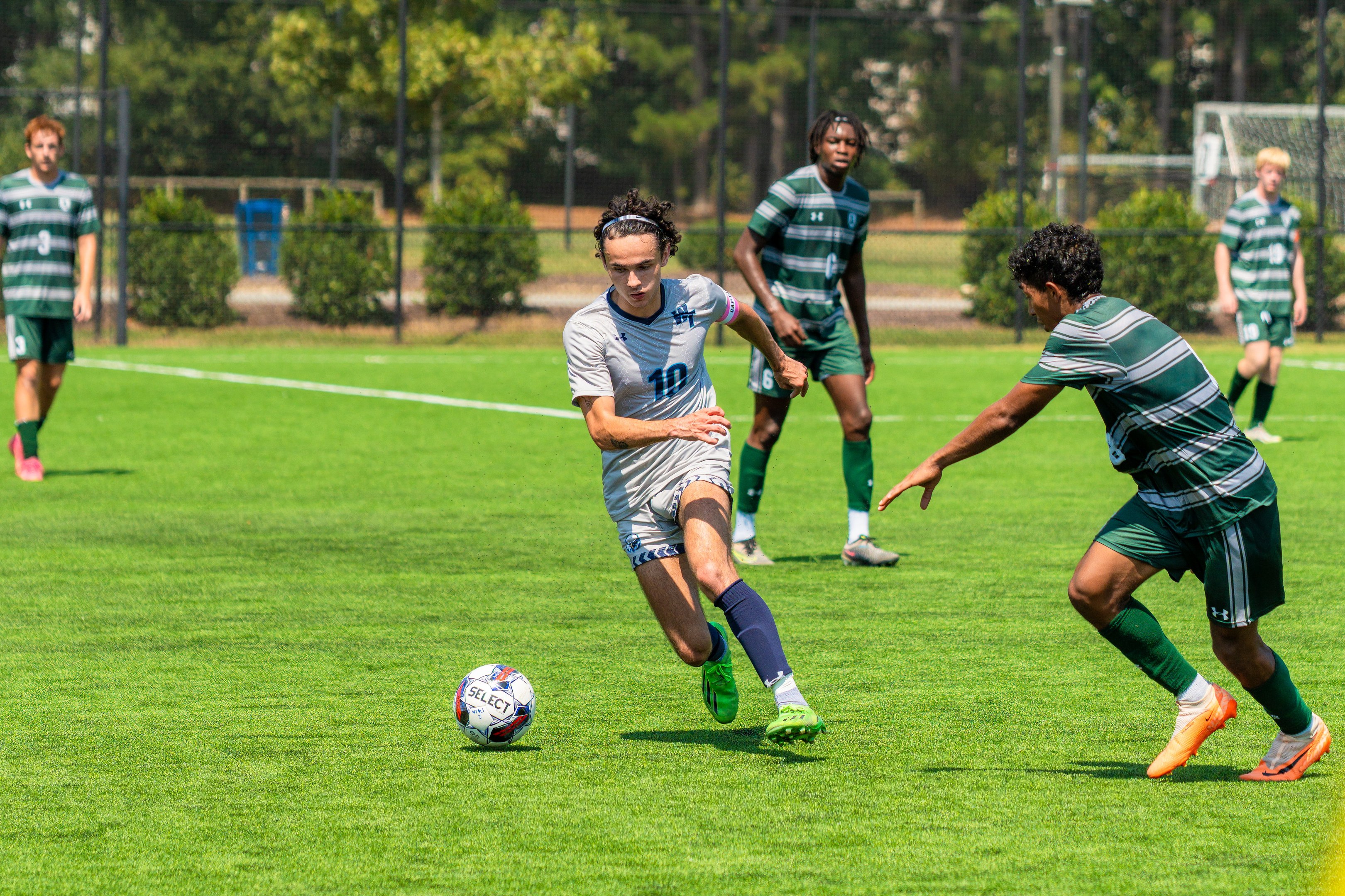 Junge Männer in Sportuniformen spielen Fußball auf einem grünen Rasen mit Bäumen, Pflanzen, Maschendraht, Pfosten und einem Tor.