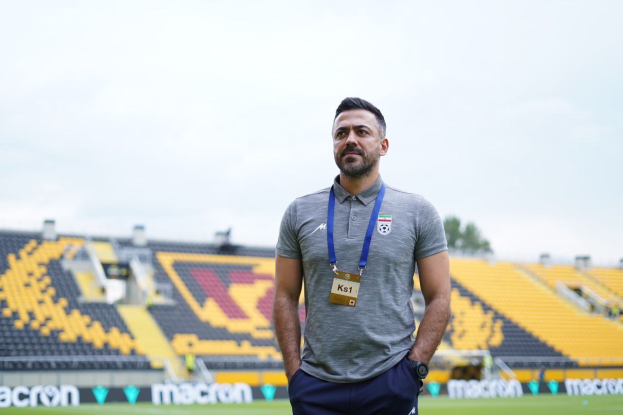 Ein als Ks1 identifizierter Mann, der Trainer der türkenischen Nationalmannschaft, steht auf einem Fußballfeld in einem grauen T-Shirt und blauen Hosen. Im Hintergrund sind gelbe Stühle, ein Baum und ein klarer blauer Himmel zu sehen.