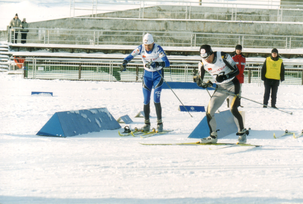 Eine Gruppe von Menschen in Winter-Sportkleidung fährt Ski auf einer schneebedeckten Piste, mit Geländern, Stufen und einer Wand im Hintergrund und Text am unteren Bildrand.