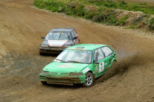 Zwei Rallye-Autos mit sichtbaren Nummern und Text auf einer staubigen Straße, umgeben von Gras, Pflanzen und Steinen.