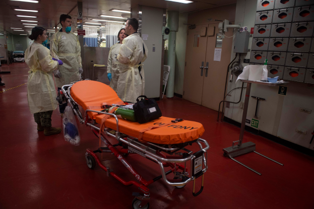 Eine Gruppe von Menschen in Schutzausrüstung steht um eine Trage in einem Krankenhauszimmer, das mit medizinischer Ausrüstung gefüllt ist, einschließlich eines Betts mit einem Beutel und anderen Gegenständen, eines Ständers mit verschiedenen Gegenständen, einer Tür, einer Säule, einer Tafel und einer Decke mit Leuchten.